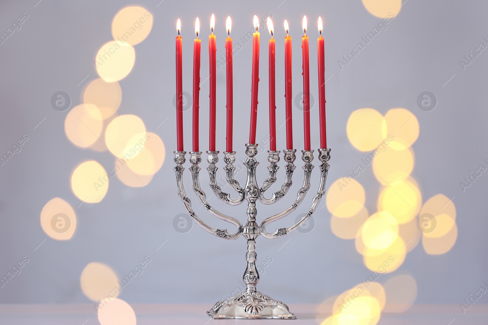 Photo of Silver menorah with burning candles against light grey background and blurred festive lights. Hanukkah celebration