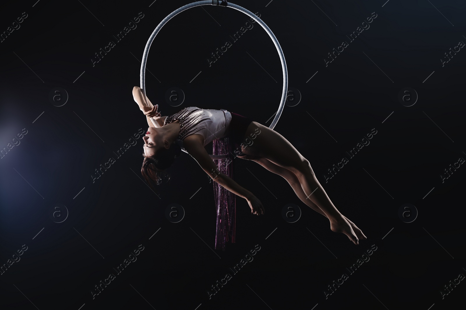 Photo of Young woman performing acrobatic element on aerial ring against dark background