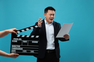 Photo of Emotional asian actor with script performing while second assistant camera holding clapperboard on light blue background. Film industry