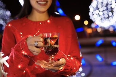 Woman with glass cup of mulled wine and garland at winter fair, closeup. Space for text