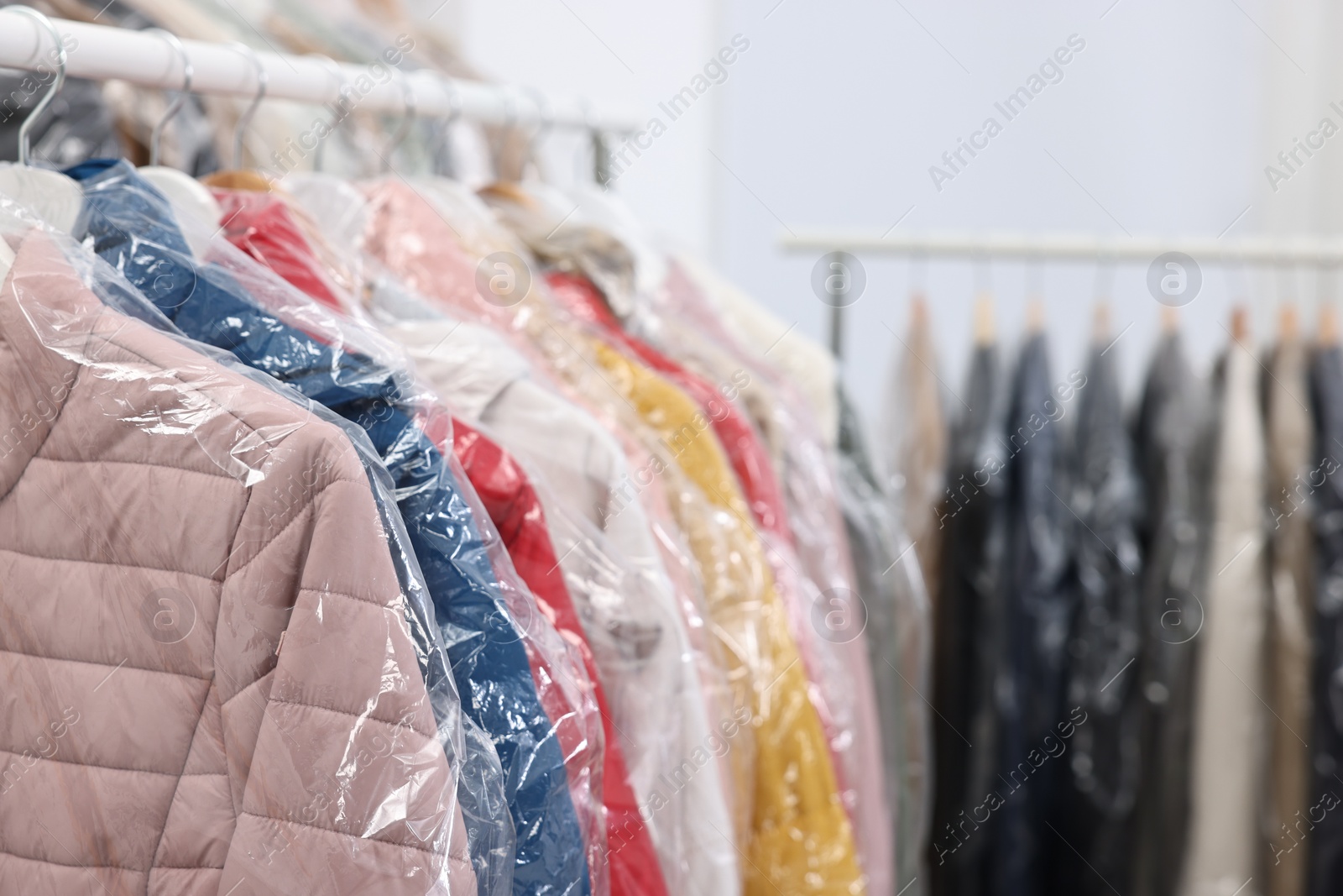 Photo of Dry-cleaning service. Many different clothes in plastic bags hanging on rack indoors, closeup