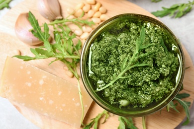 Bowl of tasty arugula pesto and ingredients on wooden board, flat lay