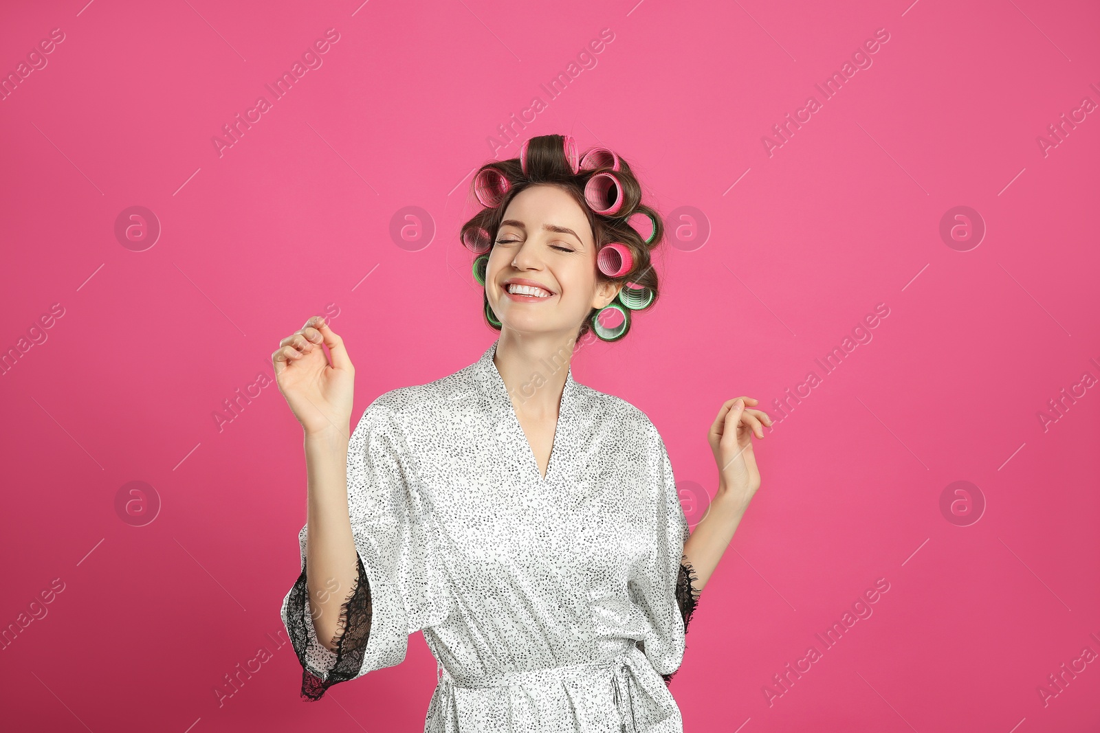 Photo of Happy young woman in silk bathrobe with hair curlers dancing on pink background