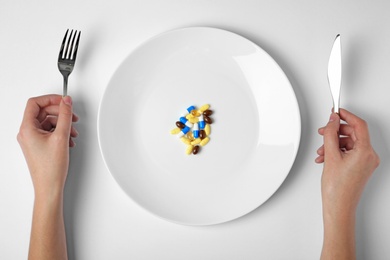 Photo of Woman holding cutlery near plate with weight loss pills on white background, top view