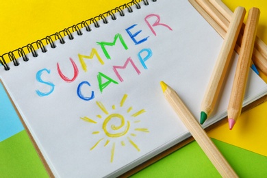 Photo of Notebook with written text SUMMER CAMP and different pencils on color background, closeup