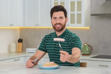 Happy man holding knife and fork with sausage at table in kitchen