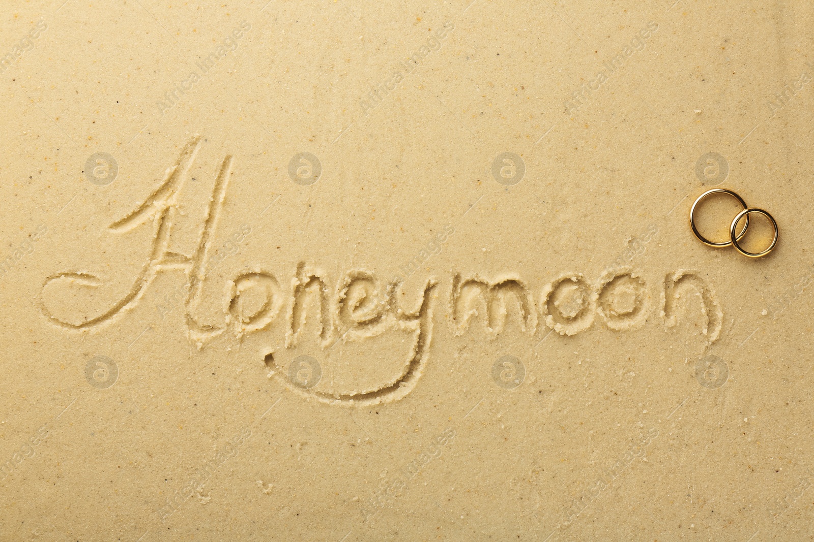 Photo of Word Honeymoon written on sand and two golden rings, top view