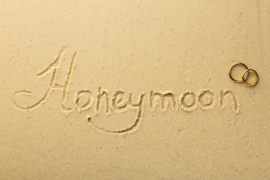 Word Honeymoon written on sand and two golden rings, top view