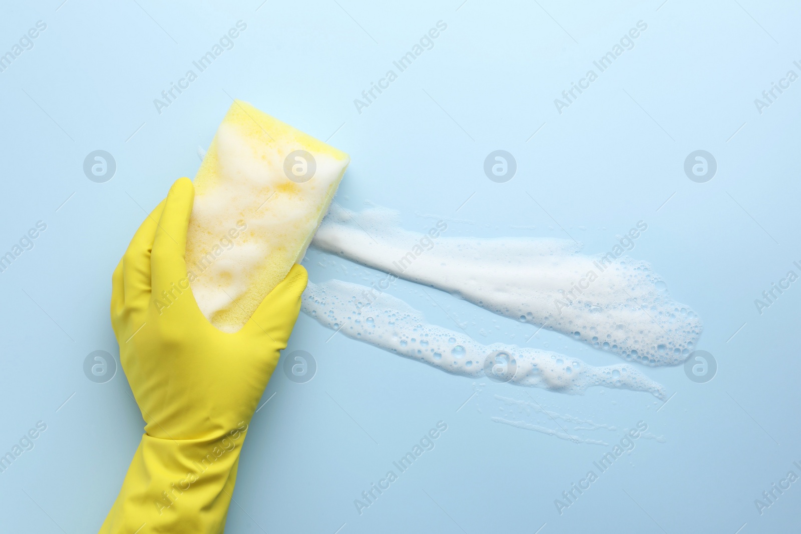 Photo of Cleaner in rubber glove holding sponge with foam on light blue background, top view