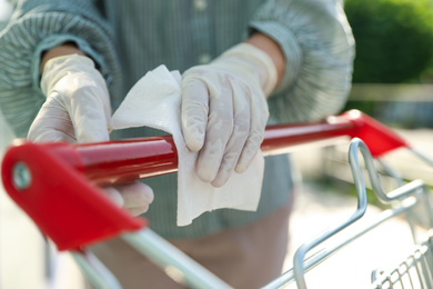 Photo of Woman cleaning handle of shopping cart with wet wipe on blurred background, closeup