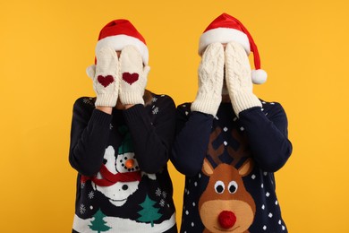 Photo of Couple in Christmas sweaters and Santa hats covering faces with hands in knitted mittens on orange background
