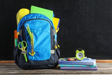 Backpack with school stationery on wooden table against blackboard