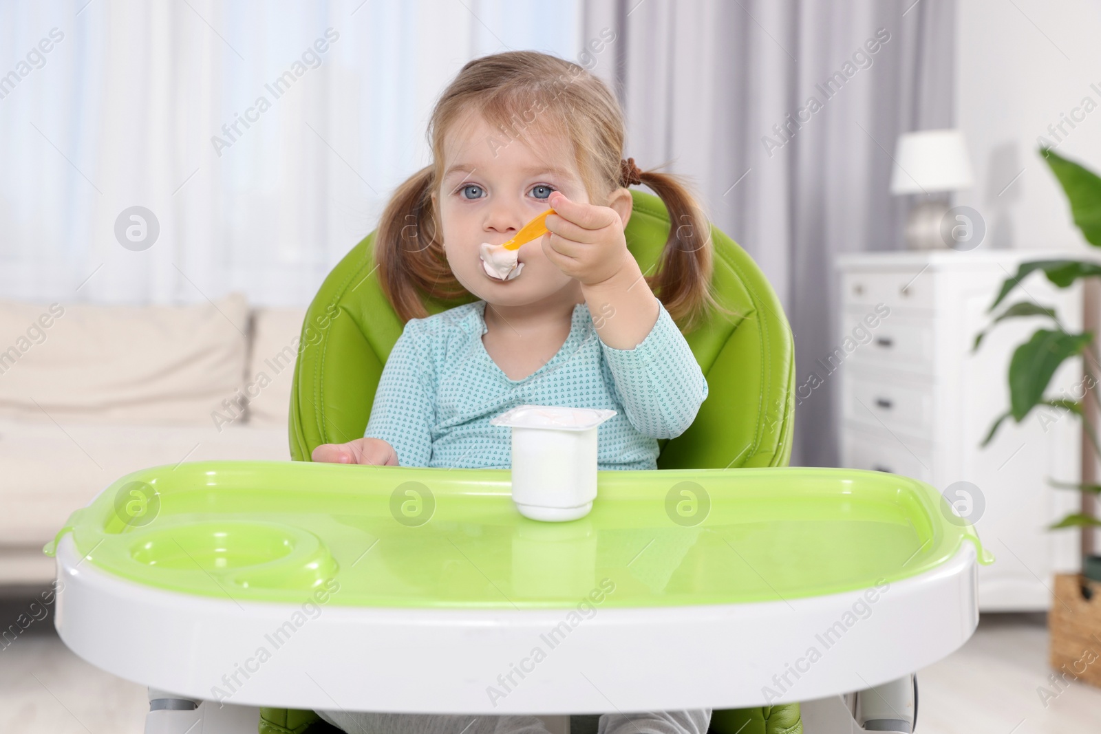 Photo of Cute little child eating tasty yogurt from plastic cup with spoon in high chair indoors