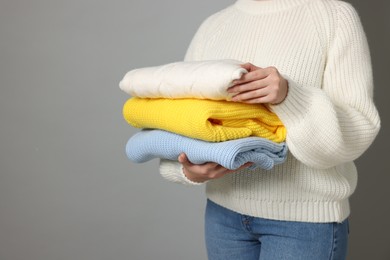 Woman with stack of knitted sweaters on grey background, closeup. Space for text
