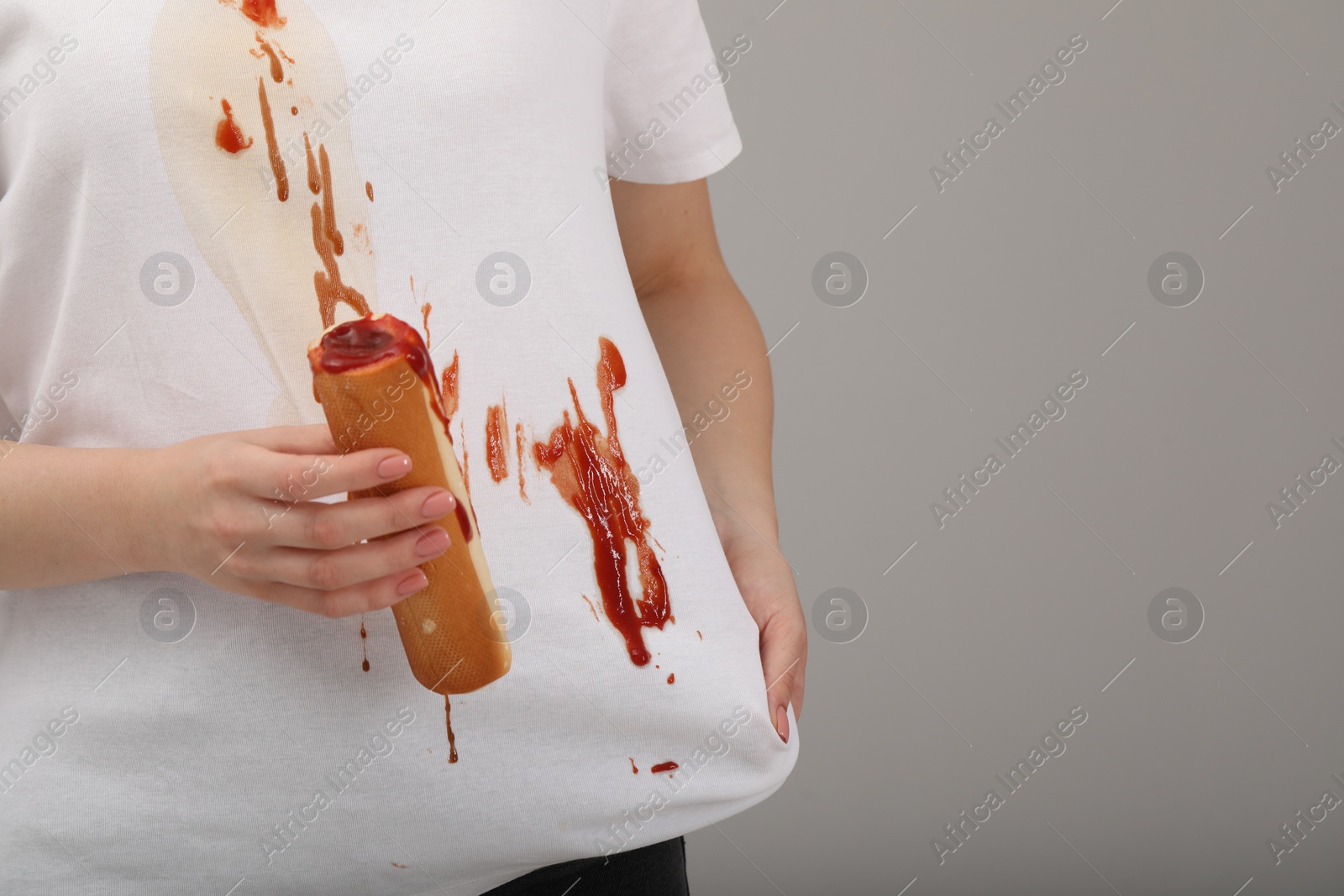 Photo of Woman holding hotdog and showing stain from sauce on her shirt against light grey background, closeup. Space for text