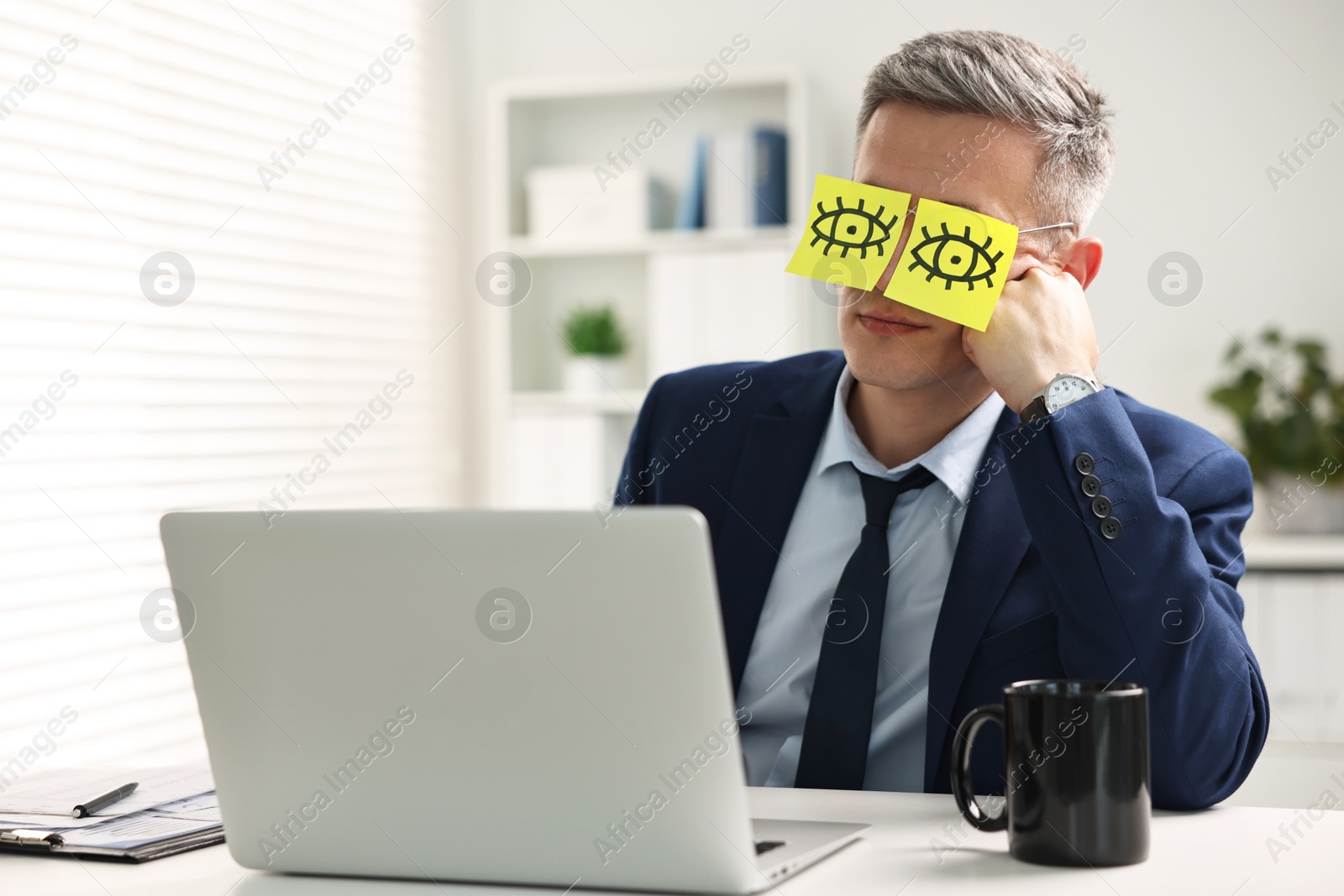 Photo of Man with fake eyes painted on sticky notes snoozing at workplace in office