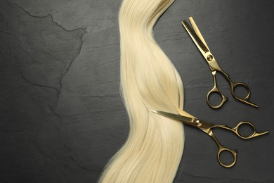Photo of Professional hairdresser scissors with blonde hair strand on dark grey table, flat lay. Space for text