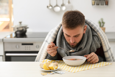 Sick young man eating tasty soup to cure flu at table in kitchen