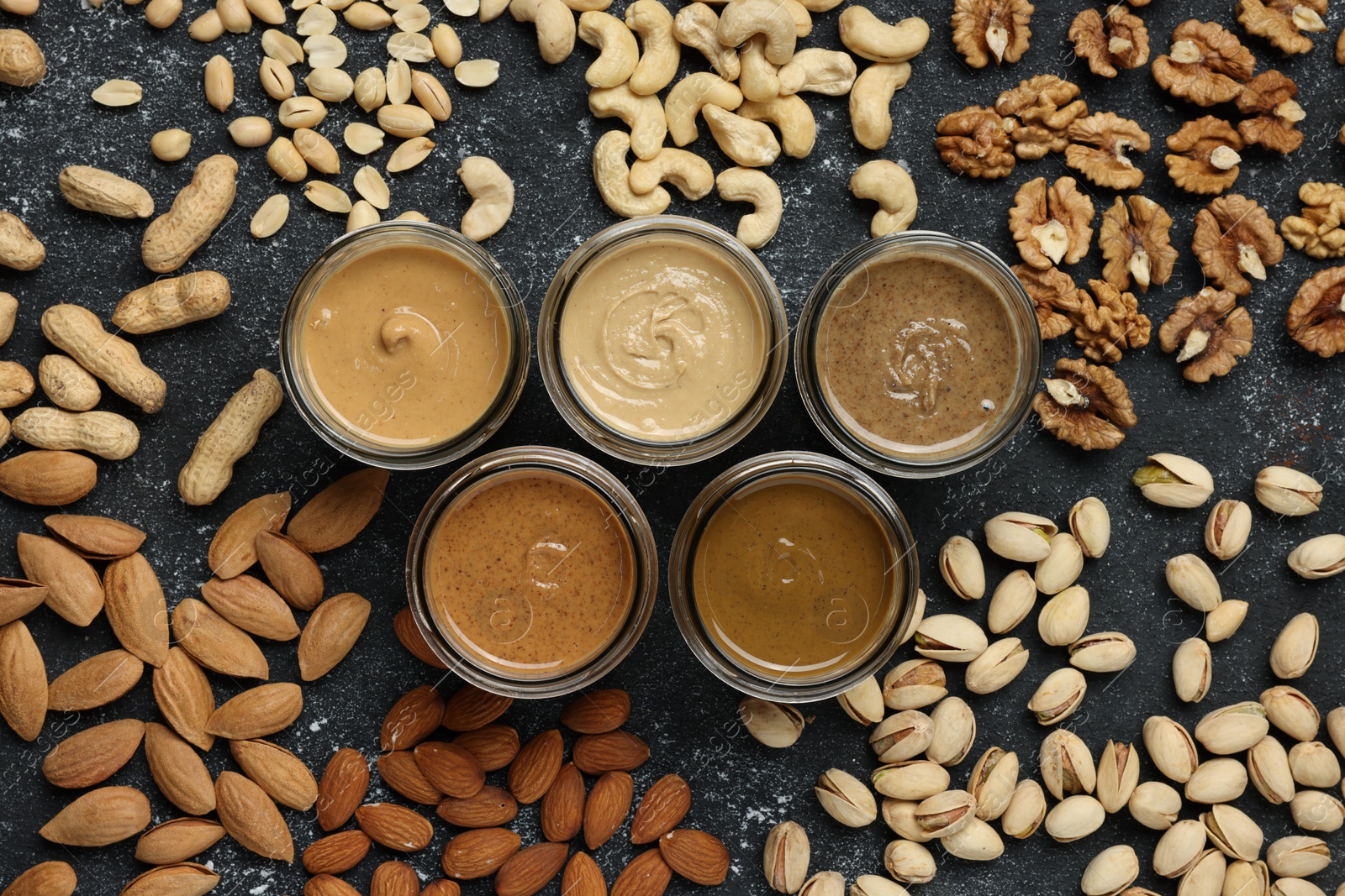 Photo of Tasty nut butters in jars and raw nuts on dark gray table, flat lay