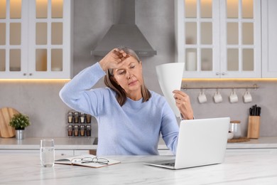 Menopause. Woman waving paper sheet to cool herself during hot flash at table in kitchen
