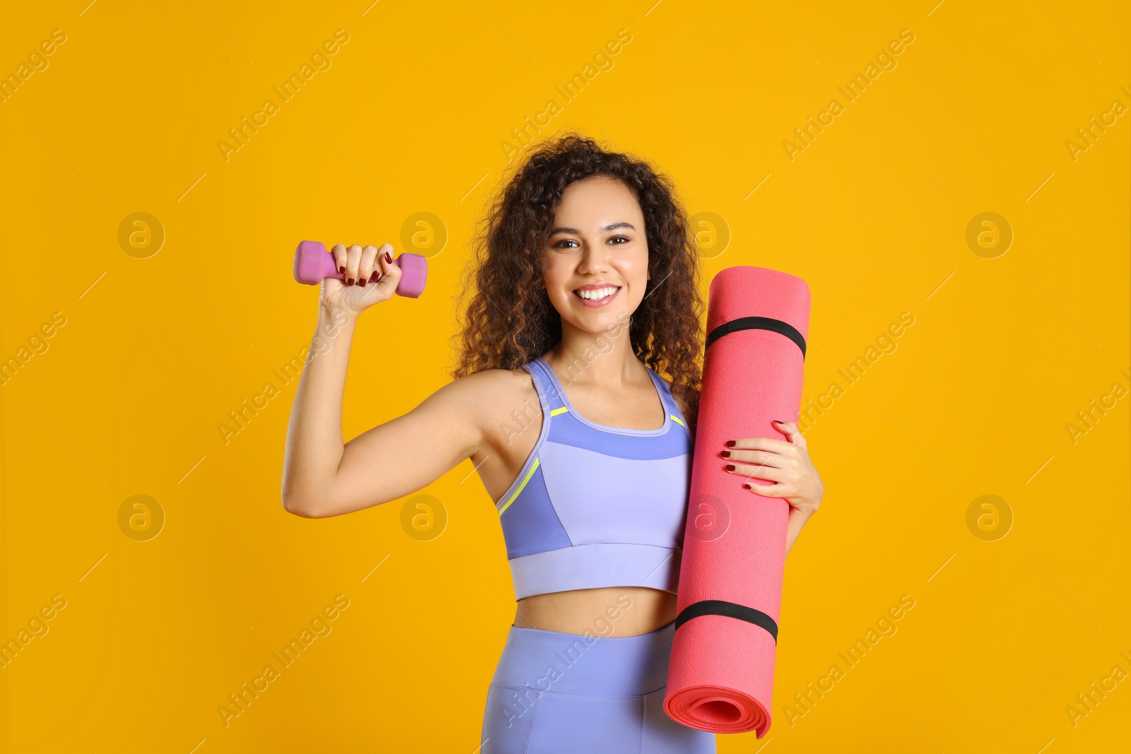 Photo of Beautiful African American woman with yoga mat and dumbbell on yellow background