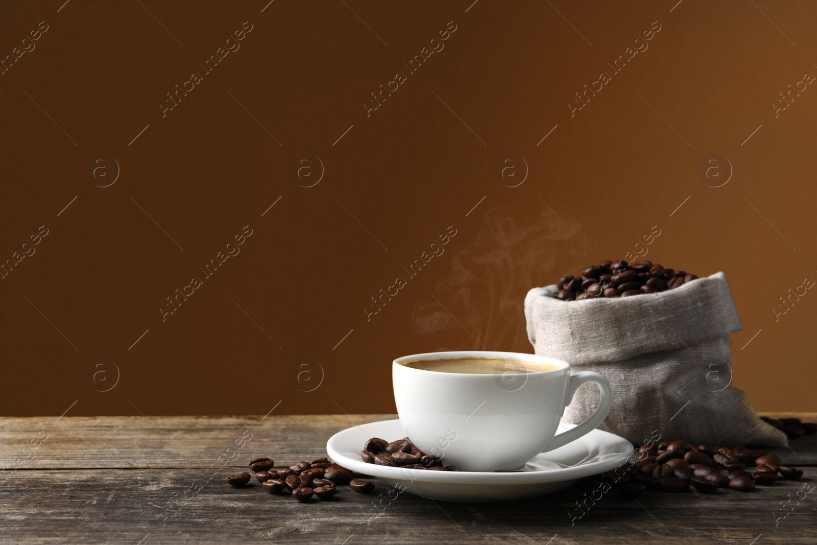 Image of Cup of hot aromatic coffee and roasted beans on wooden table against brown background. Space for text