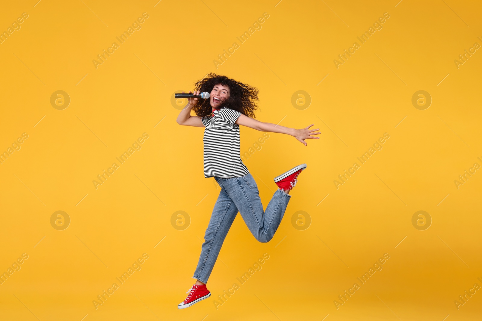 Photo of Beautiful young woman with microphone singing and jumping on yellow background