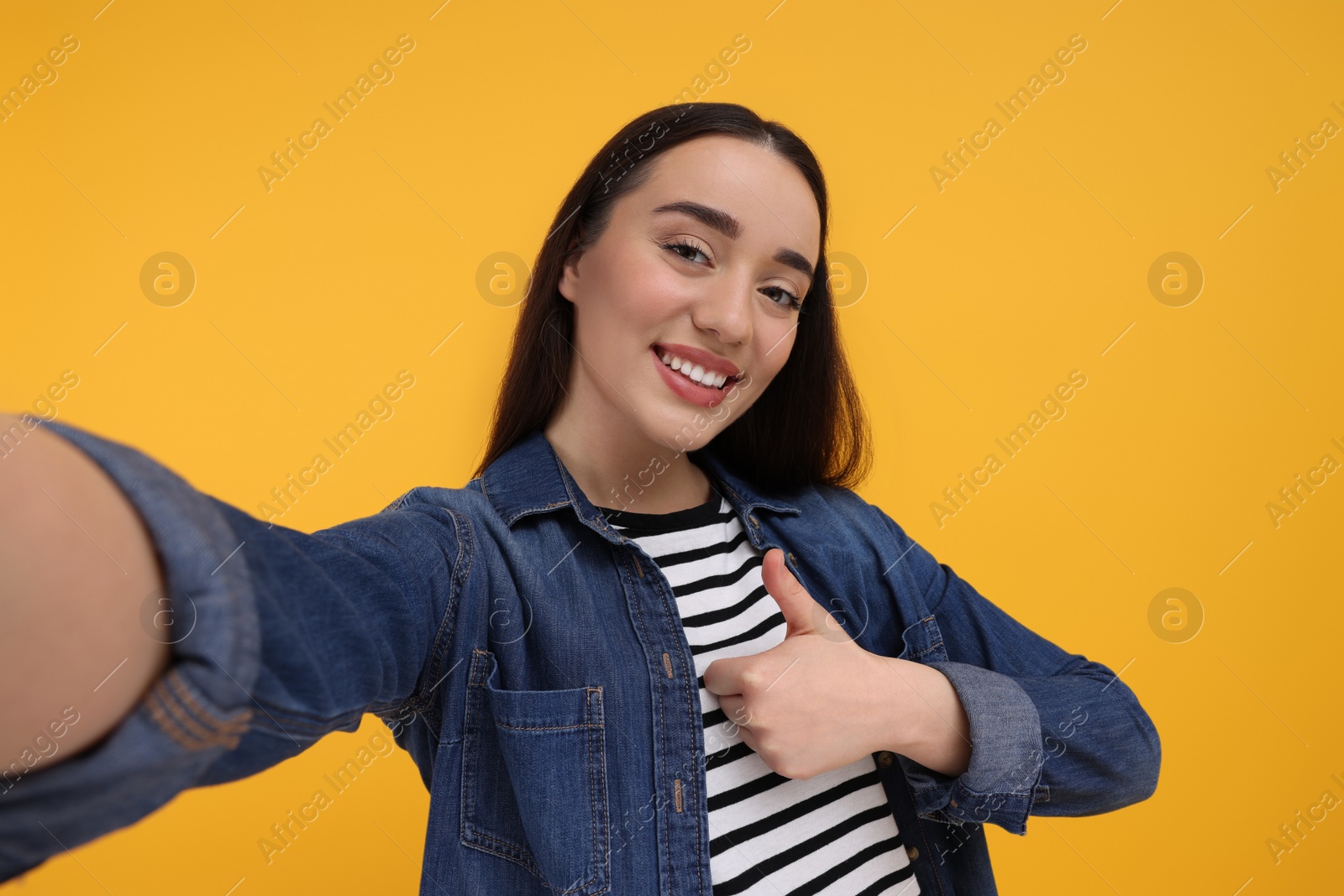 Photo of Smiling young woman taking selfie and showing thumbs up on yellow background