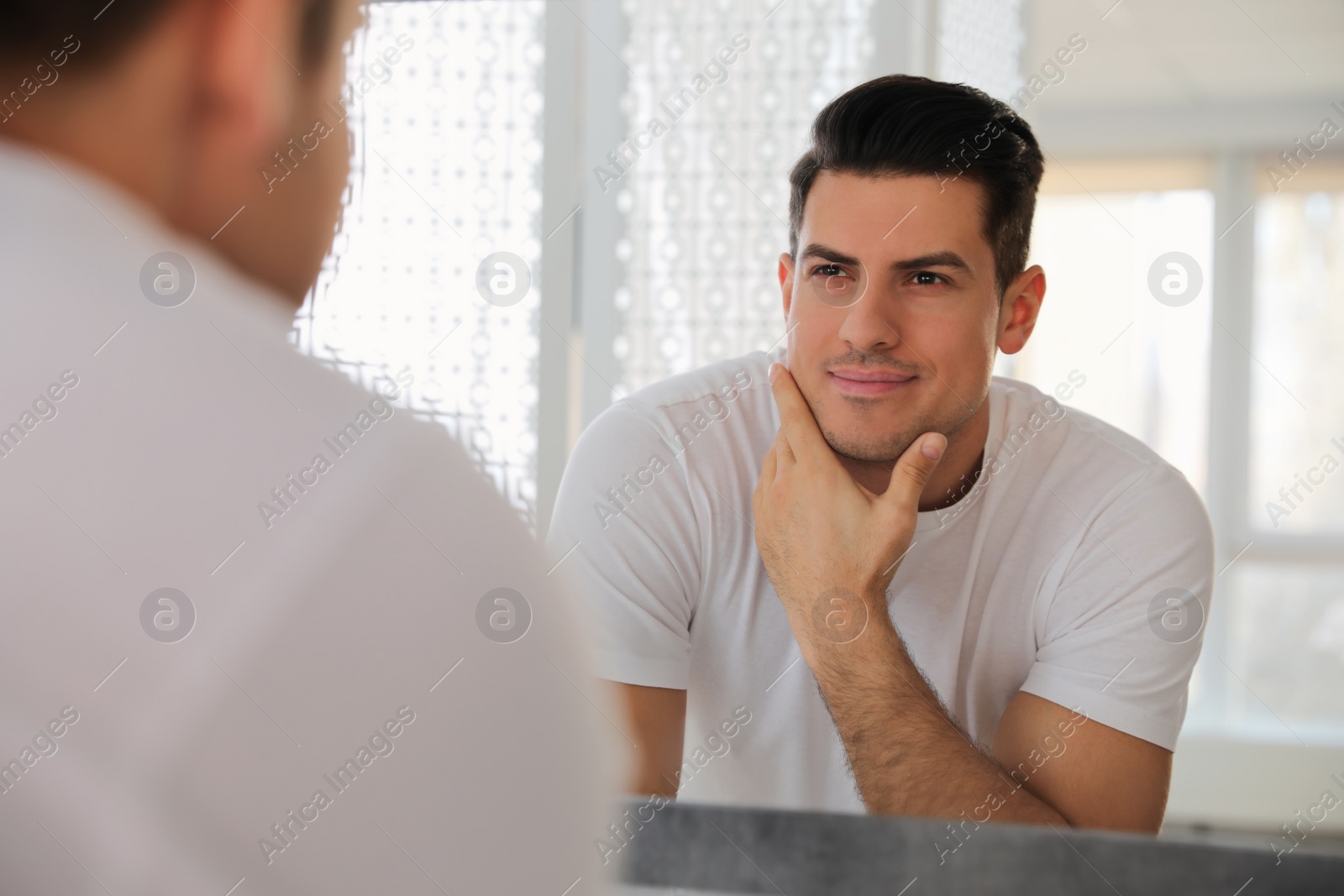 Photo of Handsome man touching his smooth face after shaving near mirror in bathroom