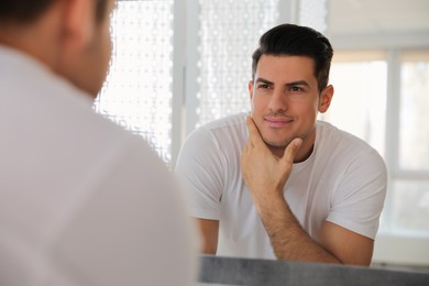 Handsome man touching his smooth face after shaving near mirror in bathroom