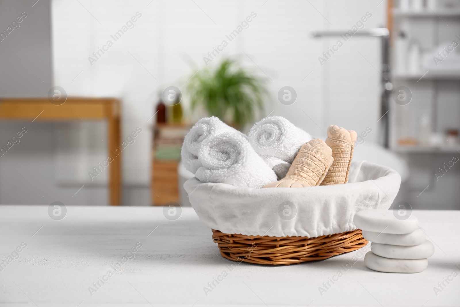 Photo of Basket with spa herbal bags, towels and stones on white table in bathroom, space for text