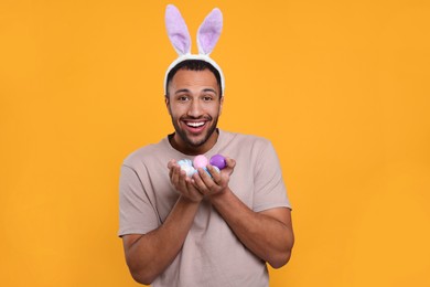 Photo of Happy African American man in bunny ears headband with Easter eggs on orange background