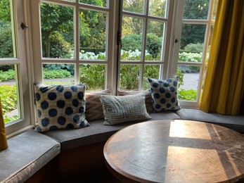 Photo of Wooden table near cozy bay window seat with cushions in room. Interior design