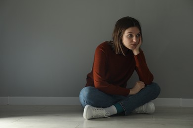 Photo of Sad young woman sitting on floor near grey wall indoors, space for text