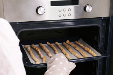 Photo of Woman taking baking sheet with homemade breadsticks out of oven, closeup. Cooking traditional grissini