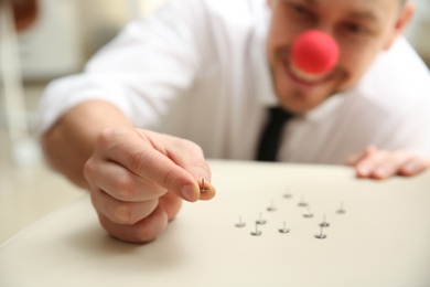 Photo of Man with clown nose putting pins onto colleague's chair in office, focus on hand. Funny joke