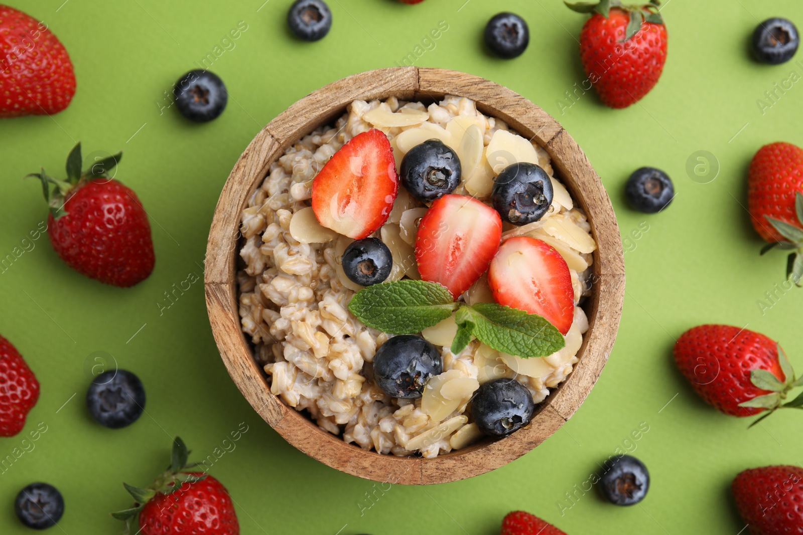 Photo of Tasty oatmeal with strawberries, blueberries and almond petals in bowl surrounded by fresh berries on green background, flat lay