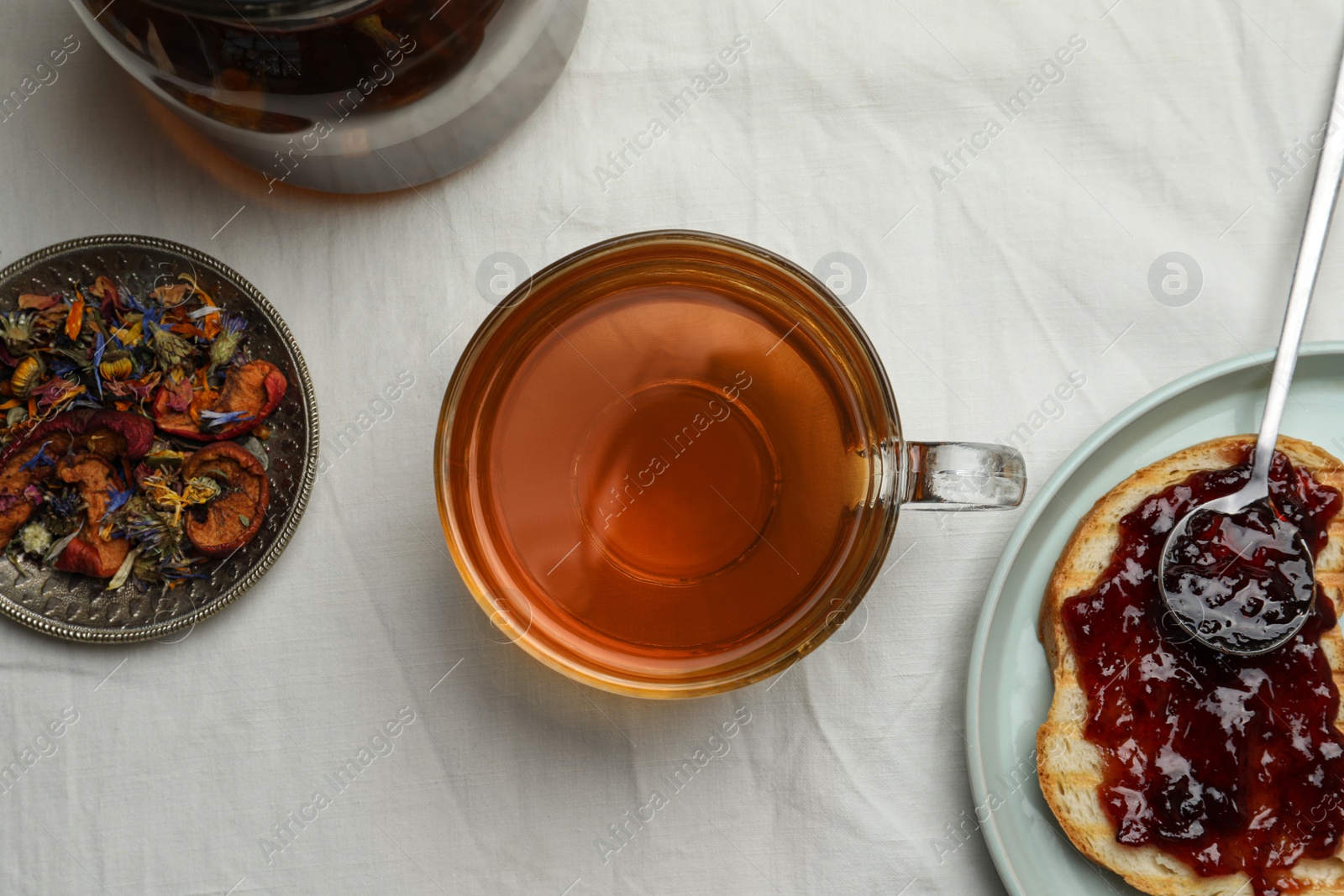 Photo of Cup of freshly brewed tea, dried herbs and bread with jam on white fabric, flat lay