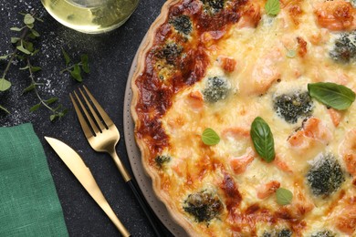 Photo of Delicious homemade salmon quiche with broccoli, basil leaves, oregano and cutlery on black table, flat lay
