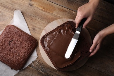 Photo of Woman smearing sponge cake with chocolate cream at wooden table, closeup. Top view