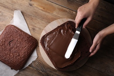 Woman smearing sponge cake with chocolate cream at wooden table, closeup. Top view