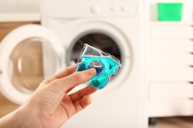 Photo of Woman holding laundry detergent capsule near washing machine indoors, closeup