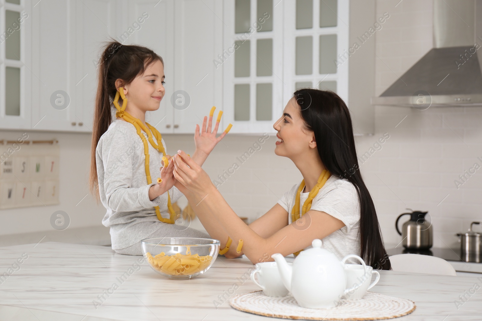 Photo of Young mother and her daughter with necklaces made of pasta having fun in kitchen