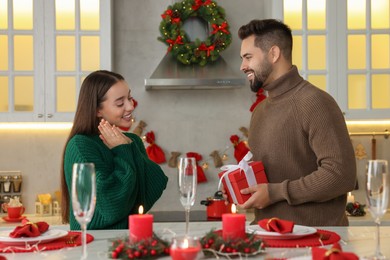 Happy young man surprising his girlfriend with Christmas gift at table in kitchen