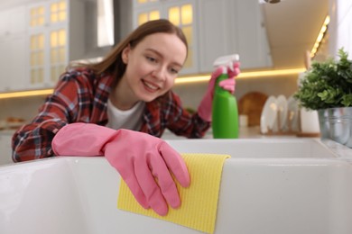 Photo of Woman with spray bottle and microfiber cloth cleaning sink in kitchen, selective focus