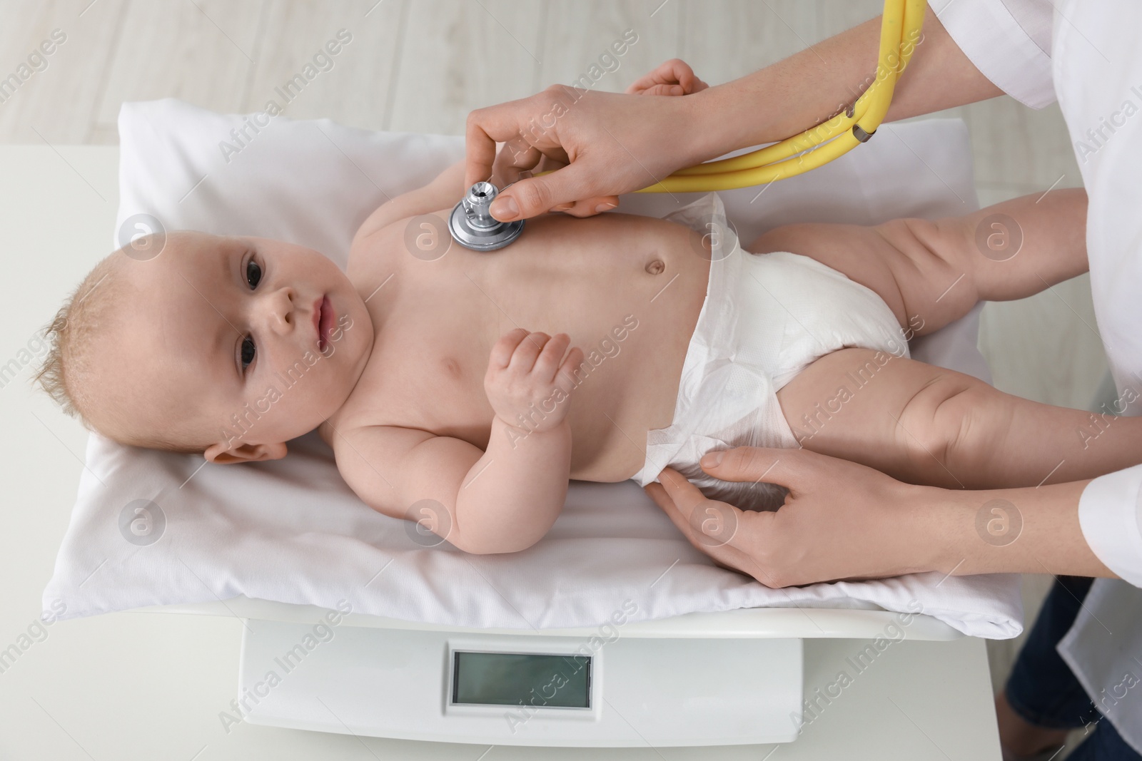 Photo of Pediatrician examining cute little baby with stethoscope and weighting it in clinic, closeup