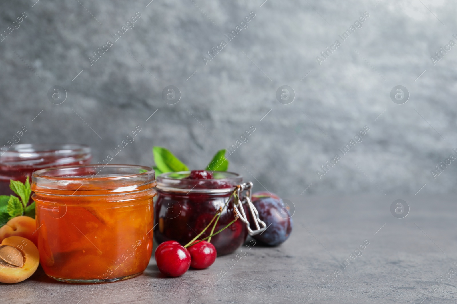 Photo of Jars with different jams and fresh fruits on grey table. Space for text