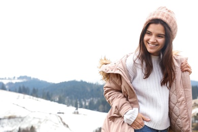 Young woman in warm clothes near snowy hill, space for text. Winter vacation