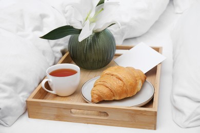 Photo of Tray with tasty croissant, cup of tea and flower on white bed