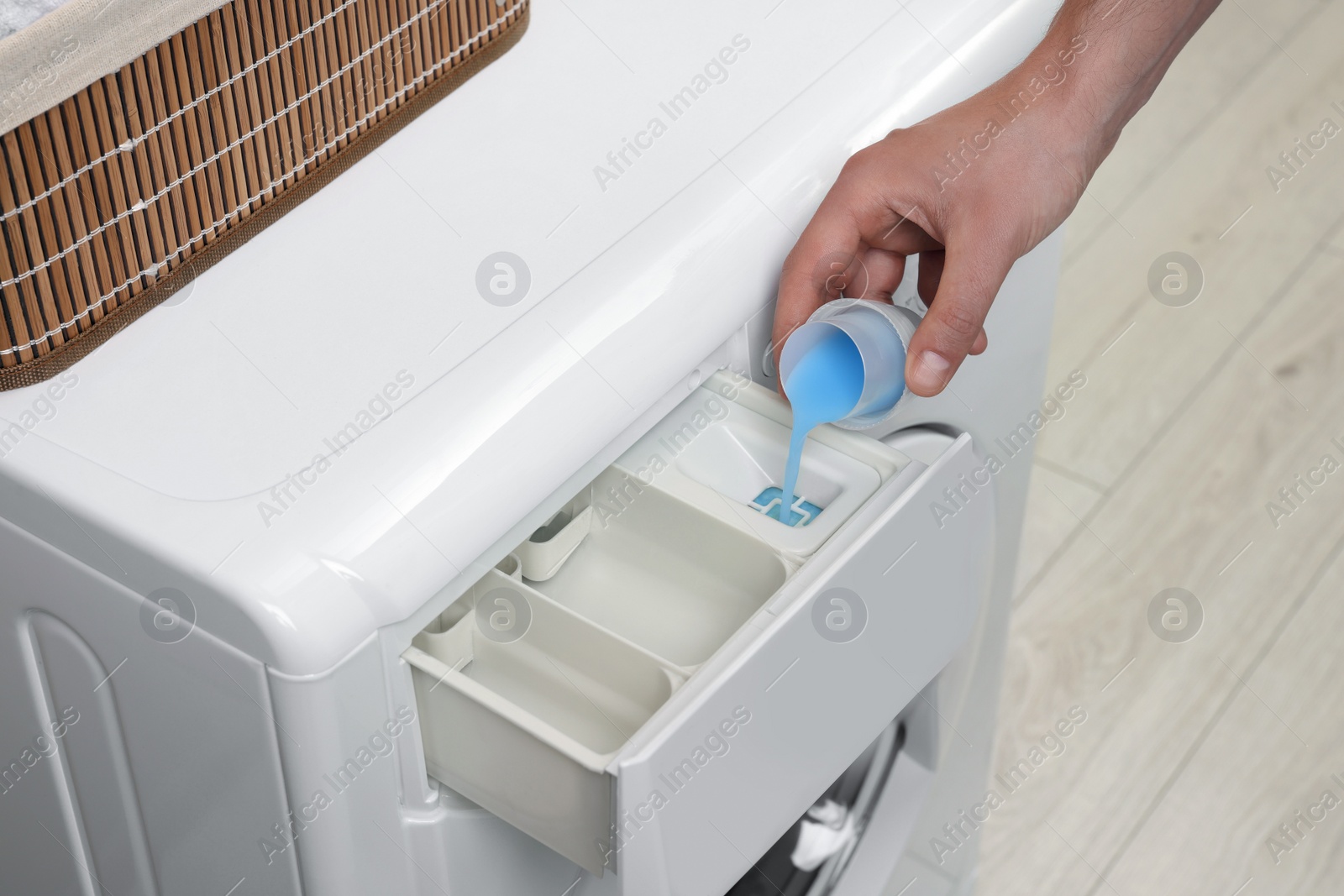Photo of Man pouring fabric softener from cap into washing machine indoors, closeup